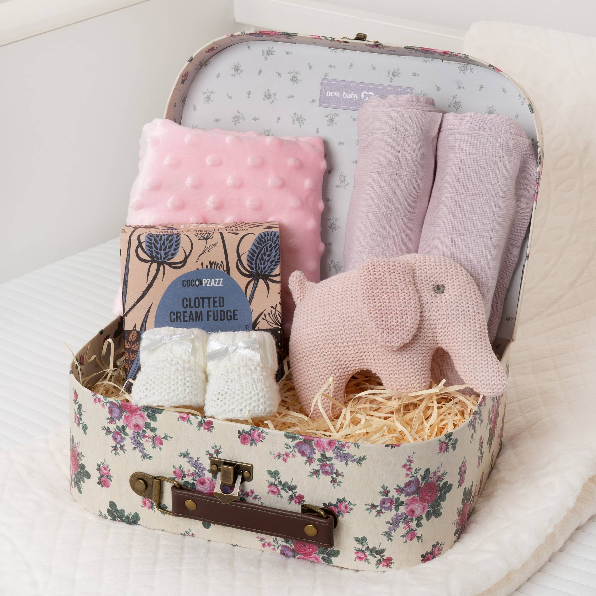 Floral-patterned suitcase filled with baby items including a pink plush elephant, pink blanket, and white fudge.