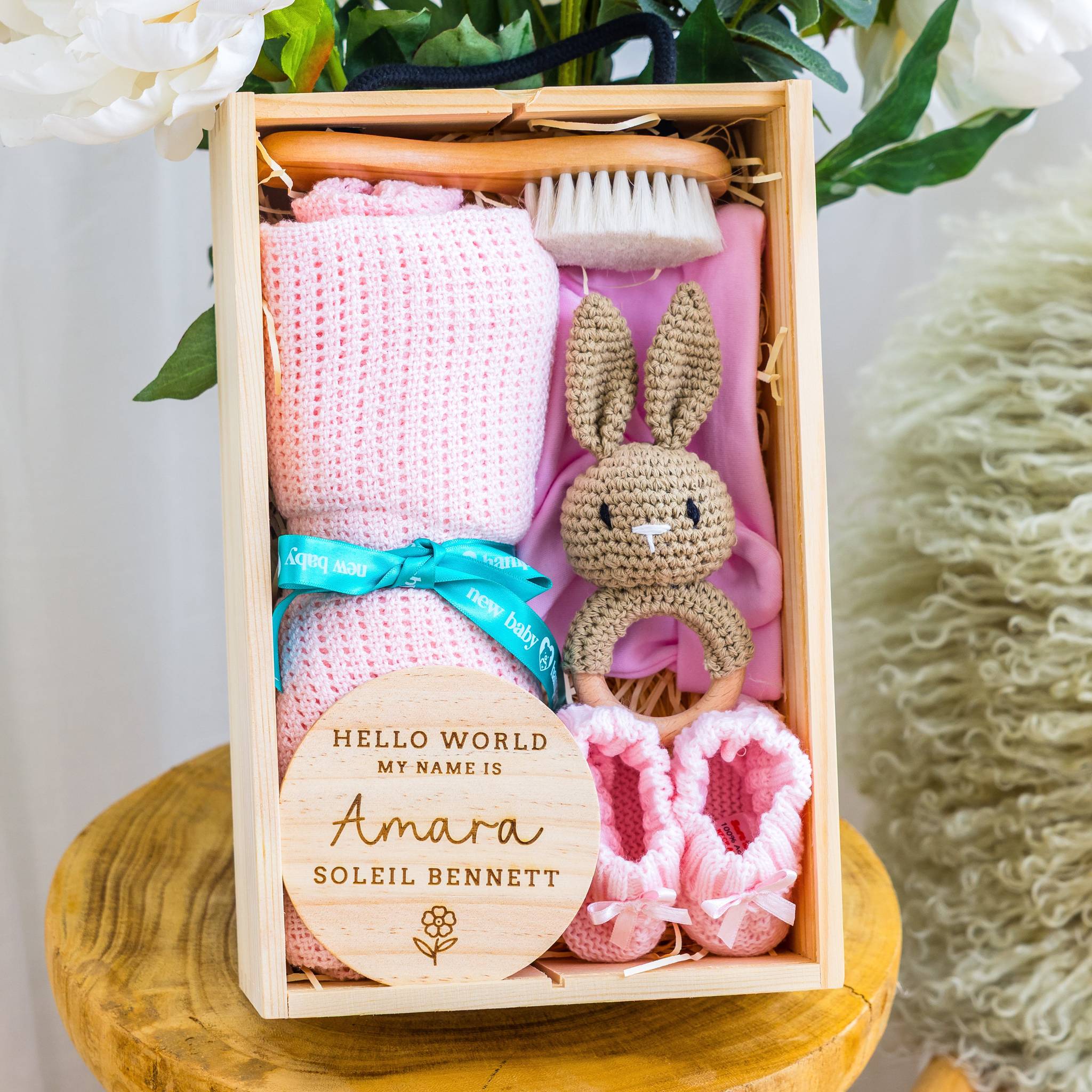 Baby gift set with pink blanket, booties, and rattle in a wooden box on a white background.